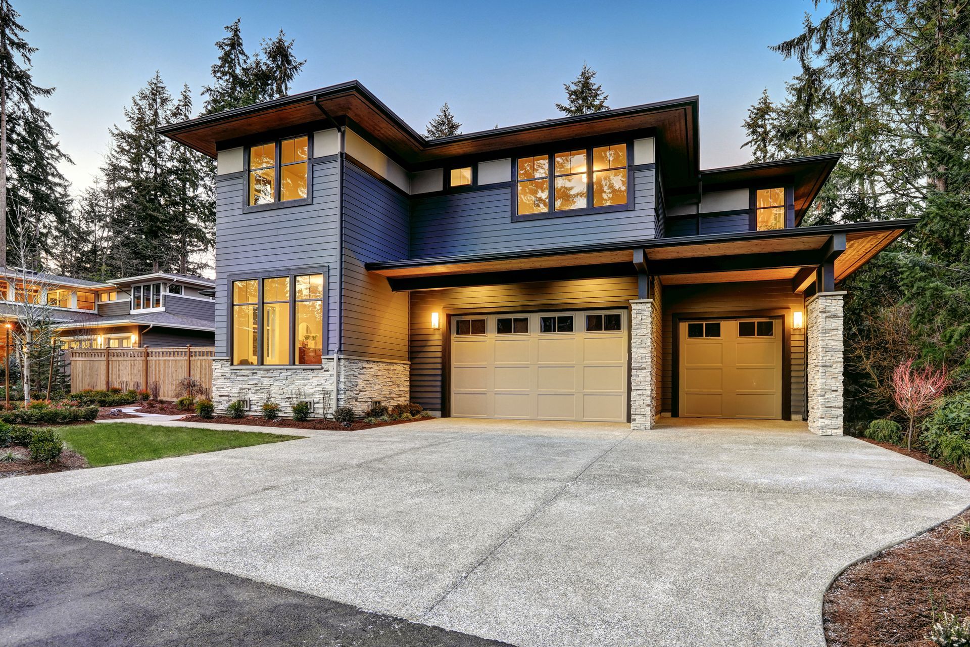 Modern two-story house with stone and dark siding, lit windows, and a three-car garage at dusk