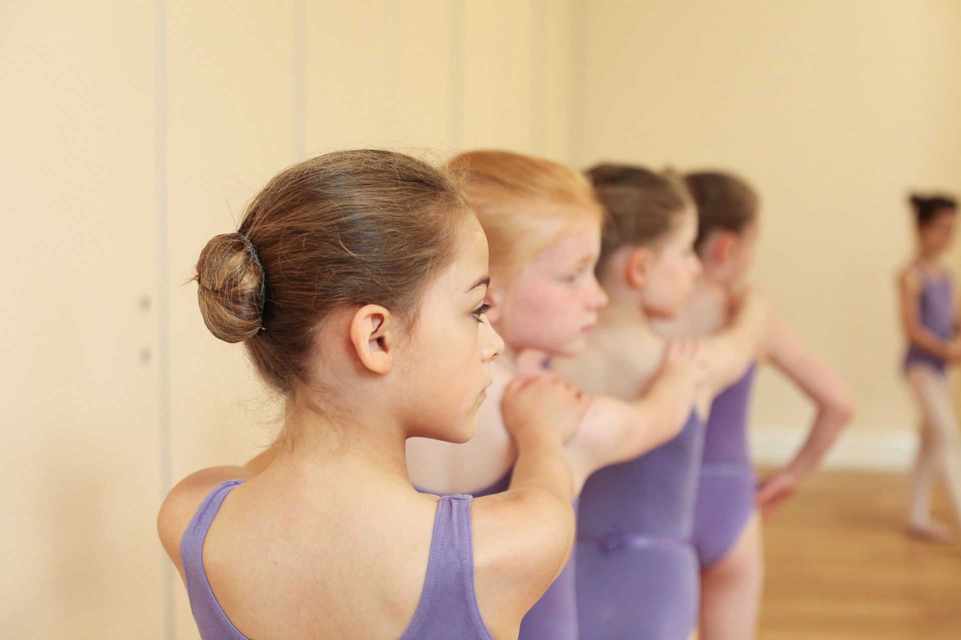 Young dancers in purple leotards stand in a line, practicing ballet in a studio.