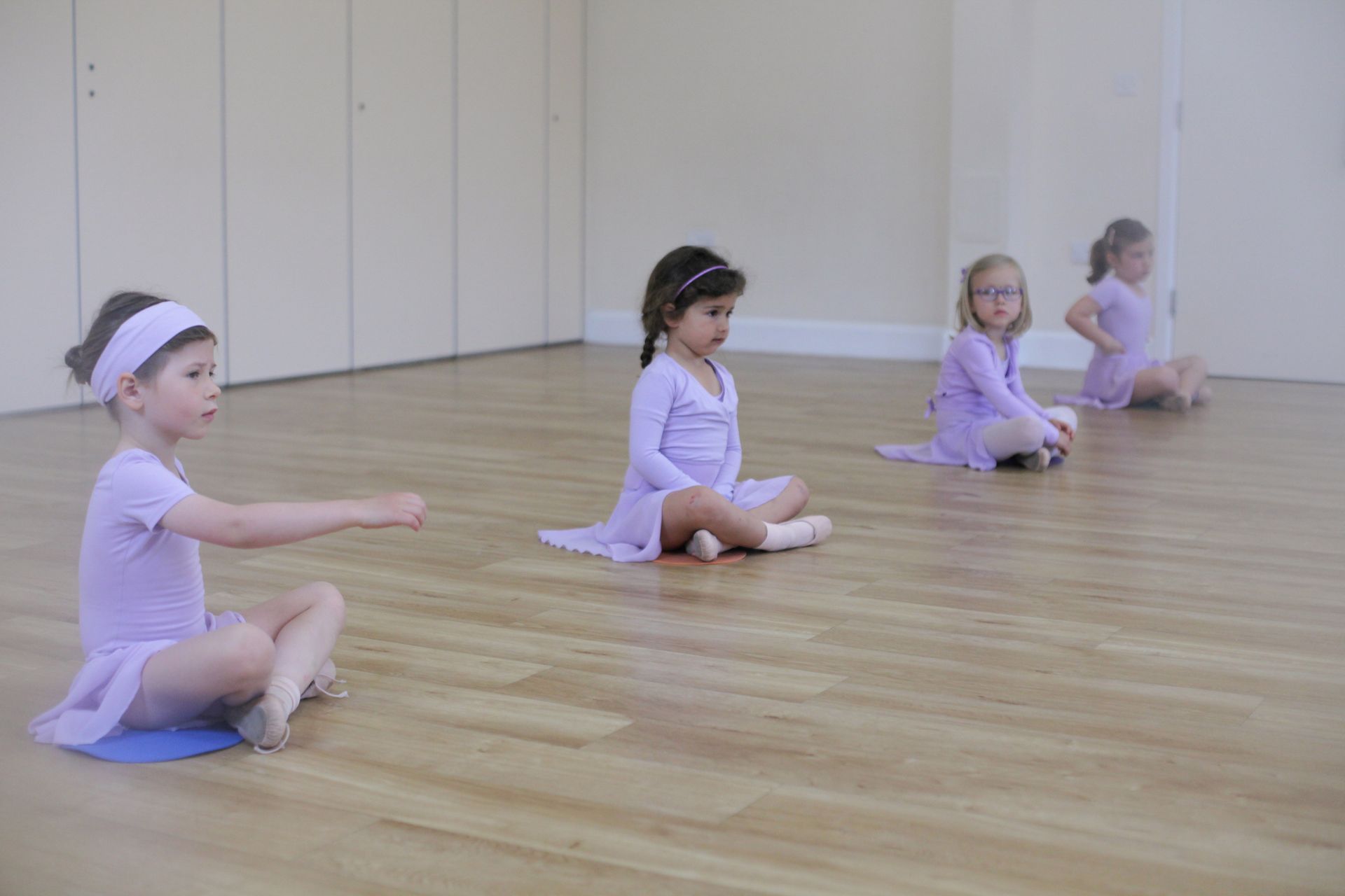 Four young children in lavender dance outfits sit on a wood floor, practicing ballet.