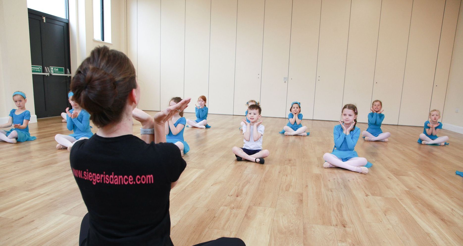 Young children sitting on the floor in a ballet class with the teacher showing dance moves.
