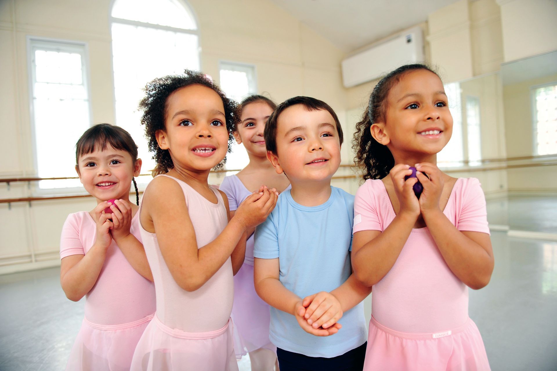 Children in a dance studio wearing pink and blue outfits, smiling and looking up.