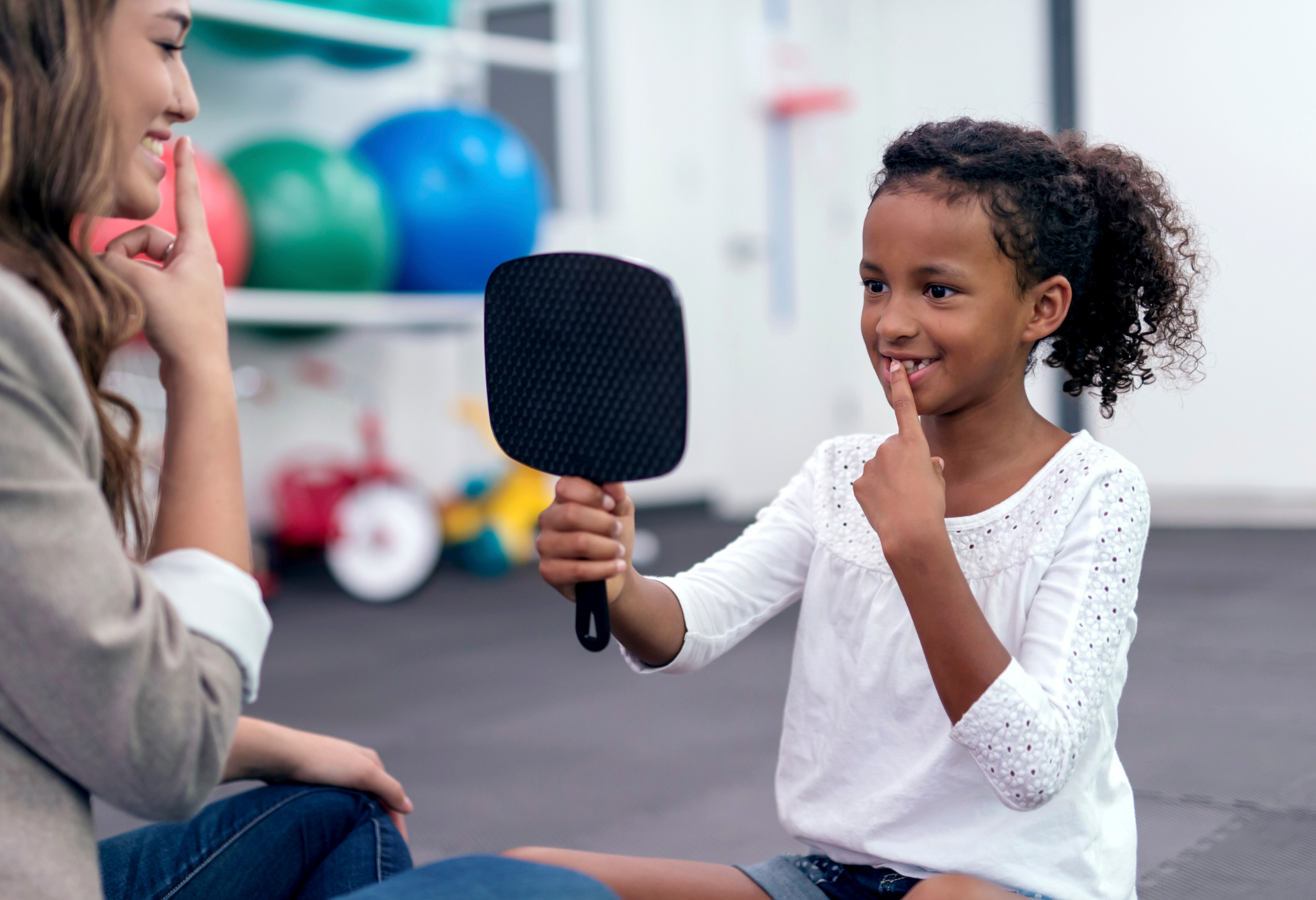 A child and adult, both pointing fingers to lips while looking into a mirror, in therapy setting.