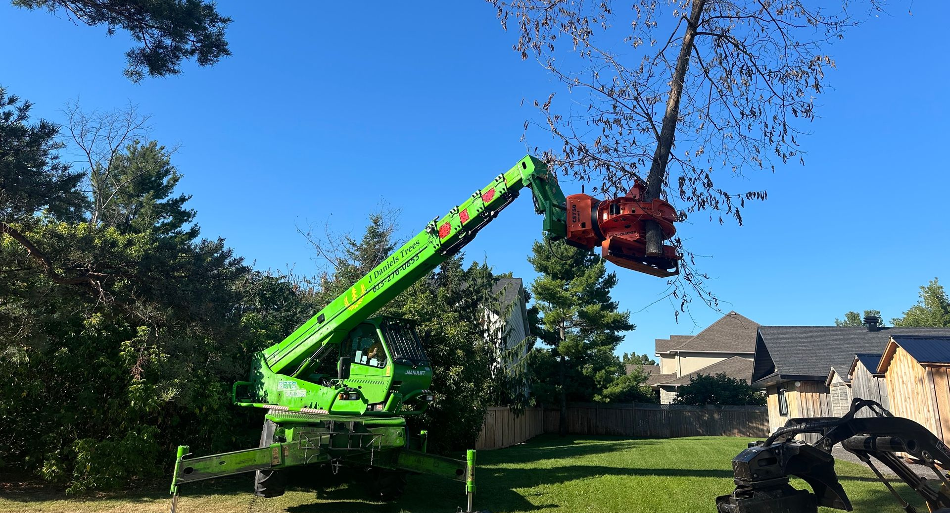A crane lifts a bare tree, with workers and equipment in a residential neighborhood under a cloudy sky.
