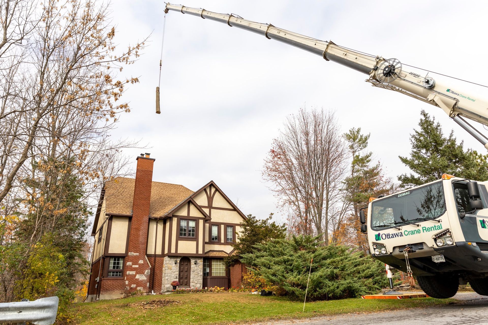 Crane lifting something over a Tudor-style house with a red brick chimney on an overcast day.