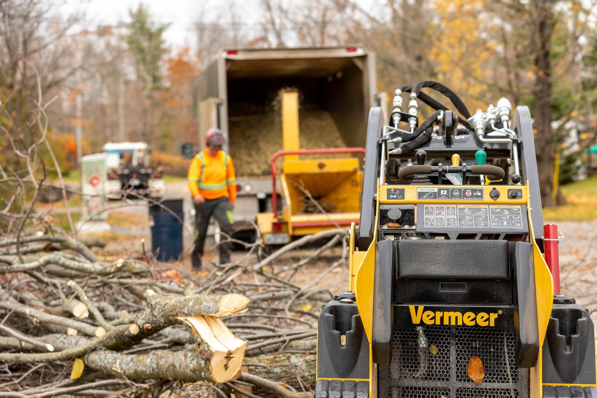 A Vermeer skid steer loader in front of a wood chipper; a worker loading the chipper with branches.