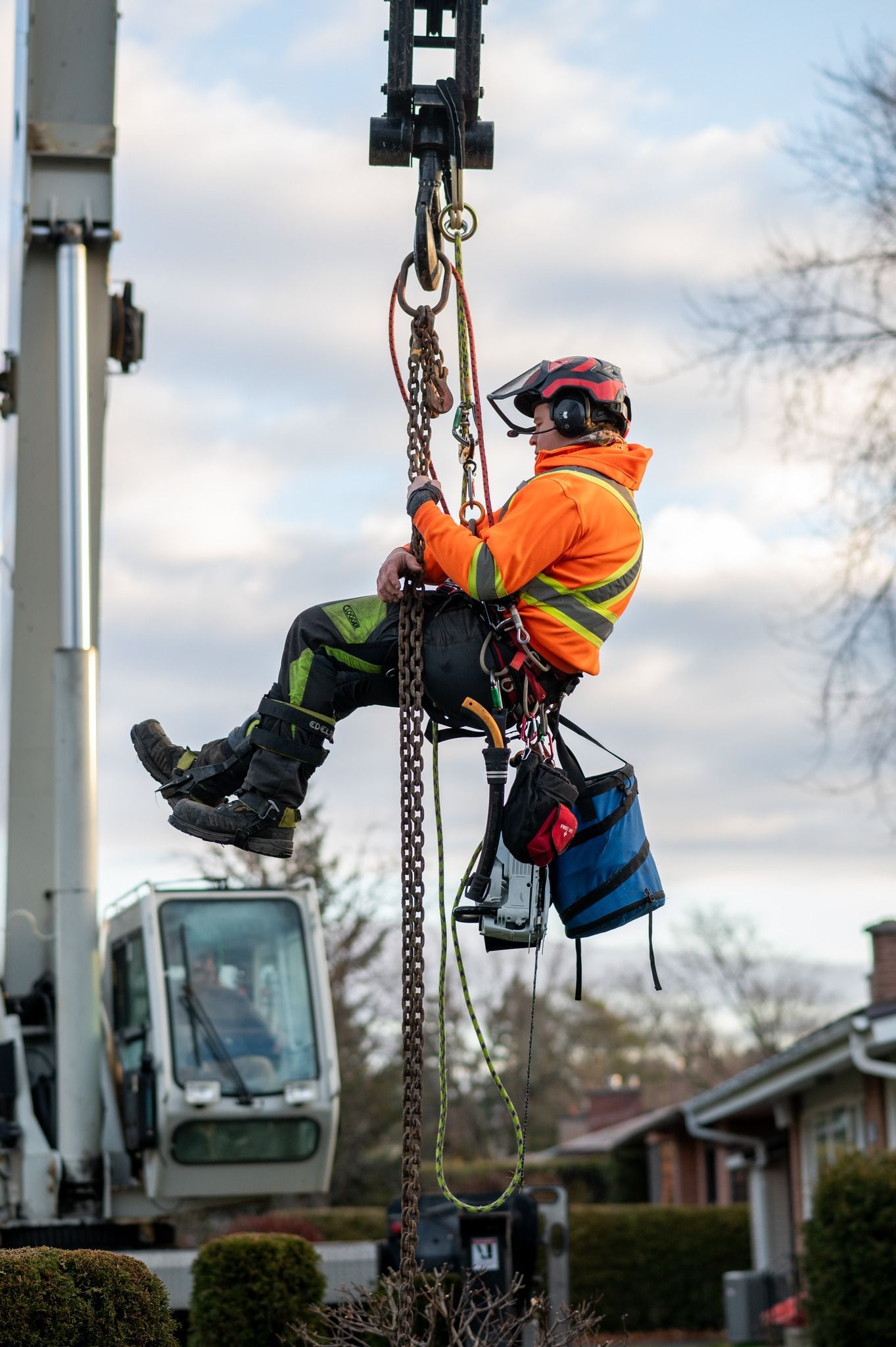 Arborist in orange jacket, helmet, and harness suspended by a crane trimming a tree. Cloudy sky, residential background.