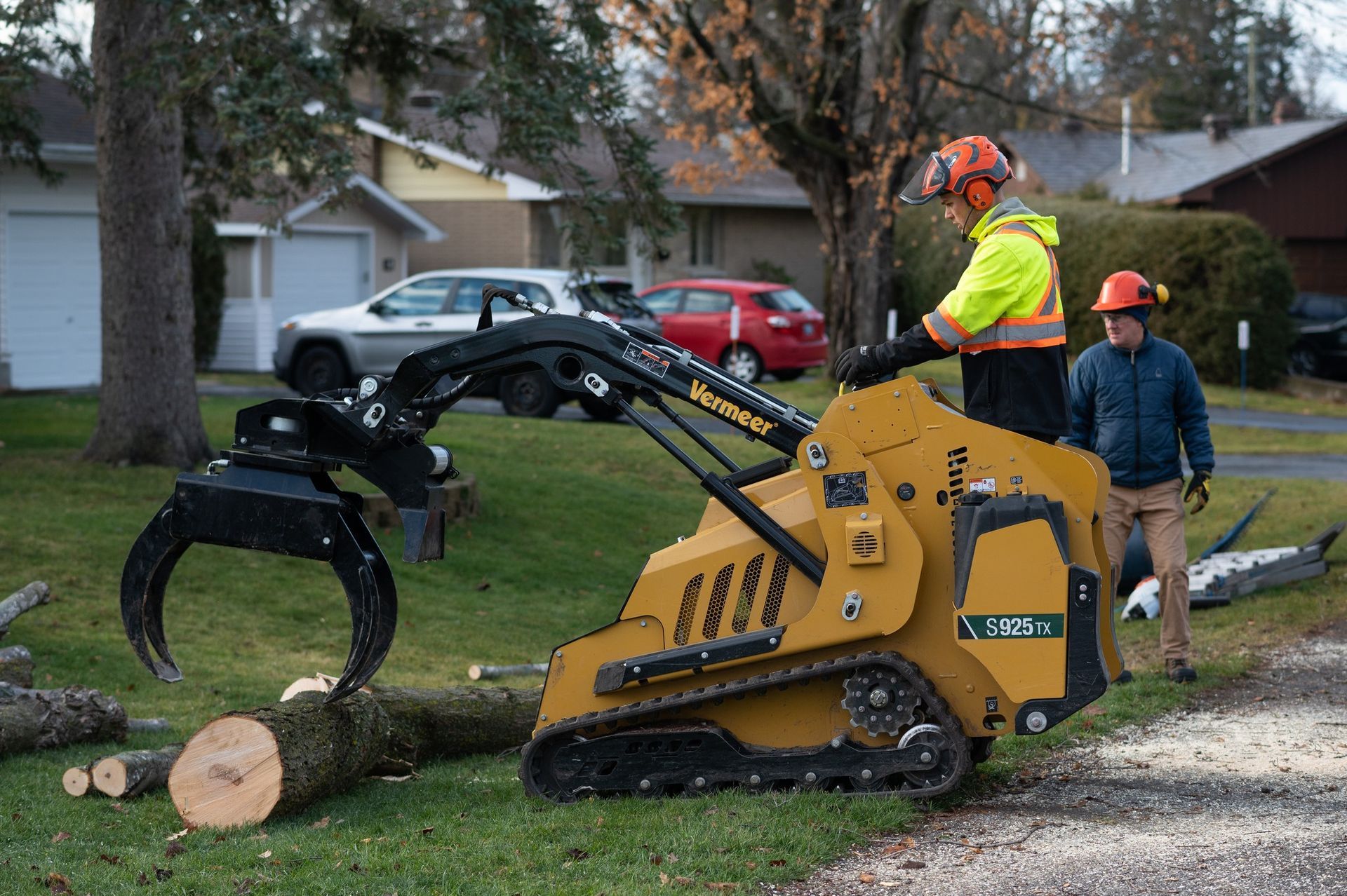 Two men using a yellow skid steer with a grapple to move logs on a grassy residential lawn.