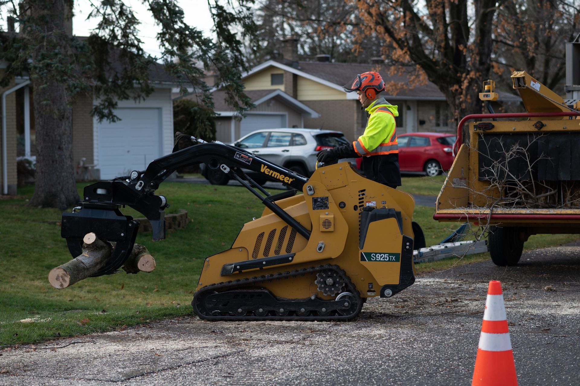 Man operating a yellow mini-skid steer with a grapple arm, chipping tree limbs on a residential street.