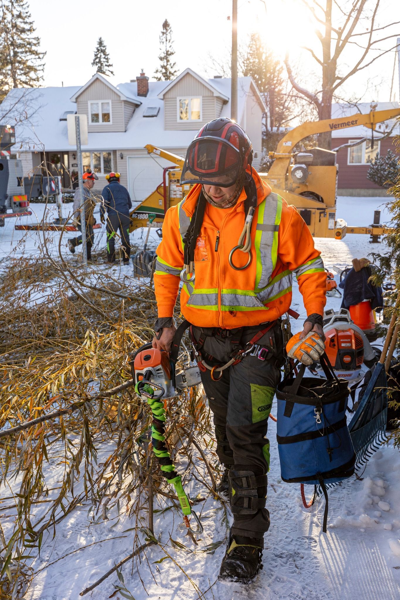Arborist in orange safety gear on snowy ground, near cut tree branches and a wood chipper.