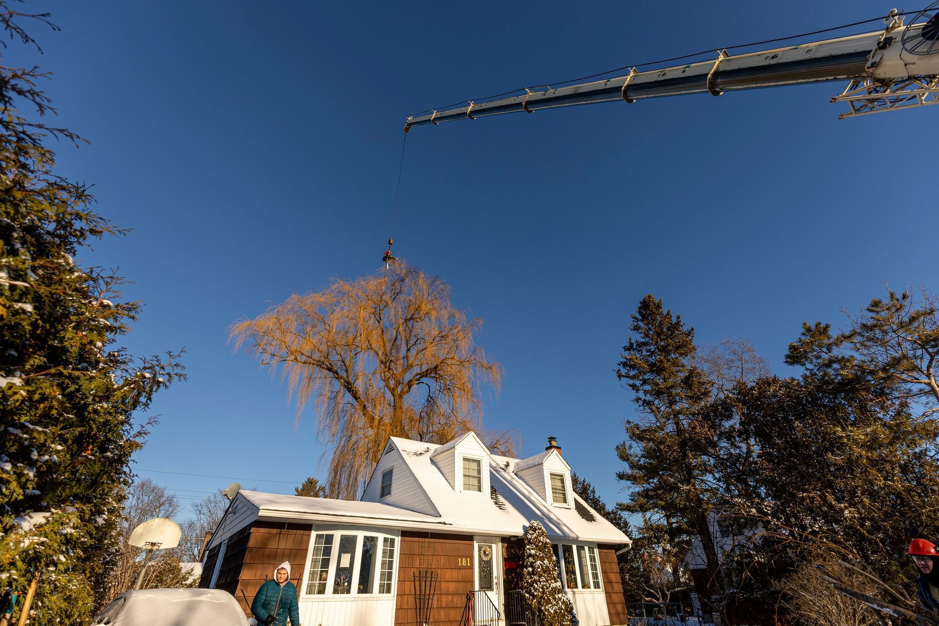 A snow-covered house with a clear blue sky and a crane overhead. Person standing by the house.