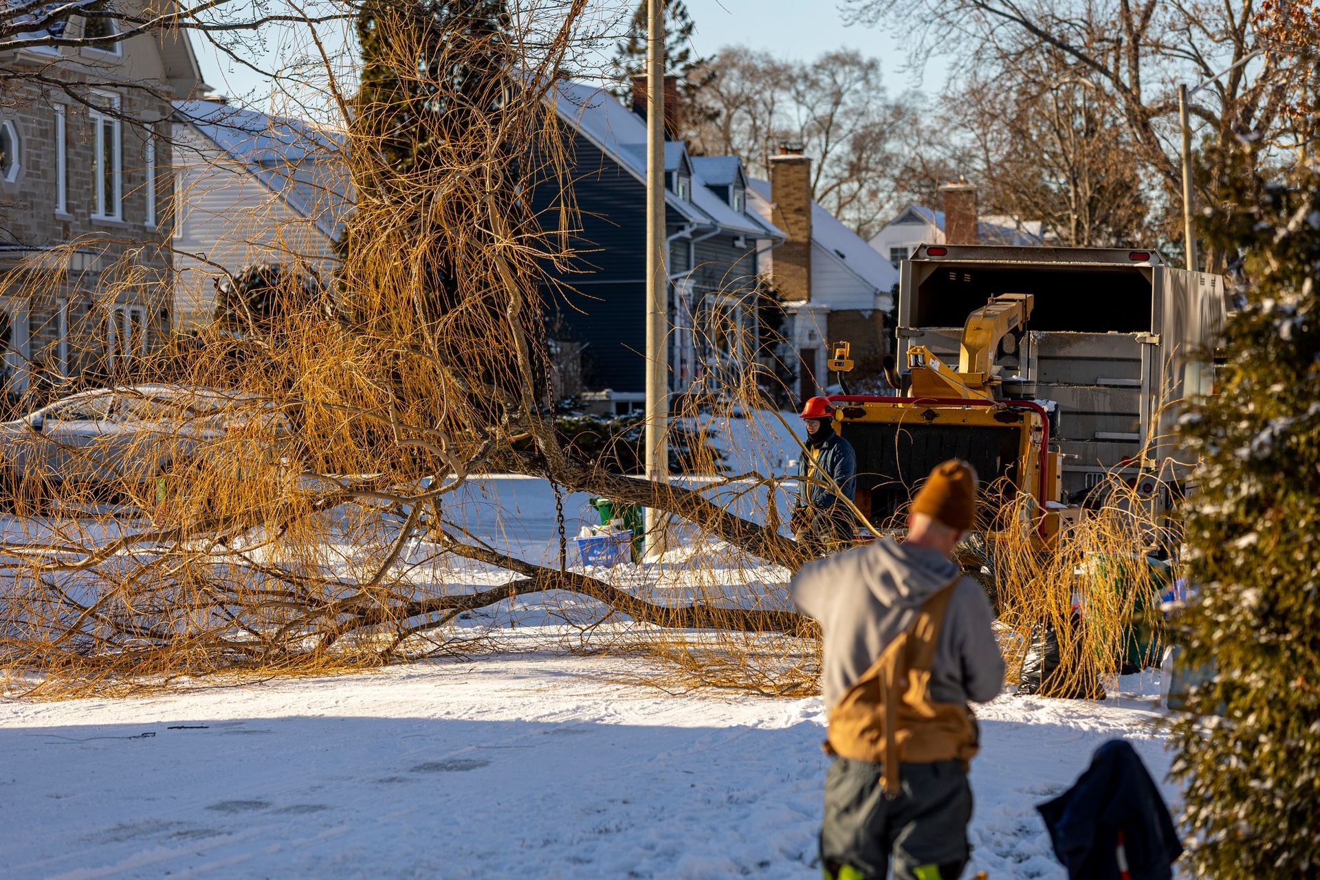 Men removing tree branches from a snowy street with a wood chipper and truck in a residential area.