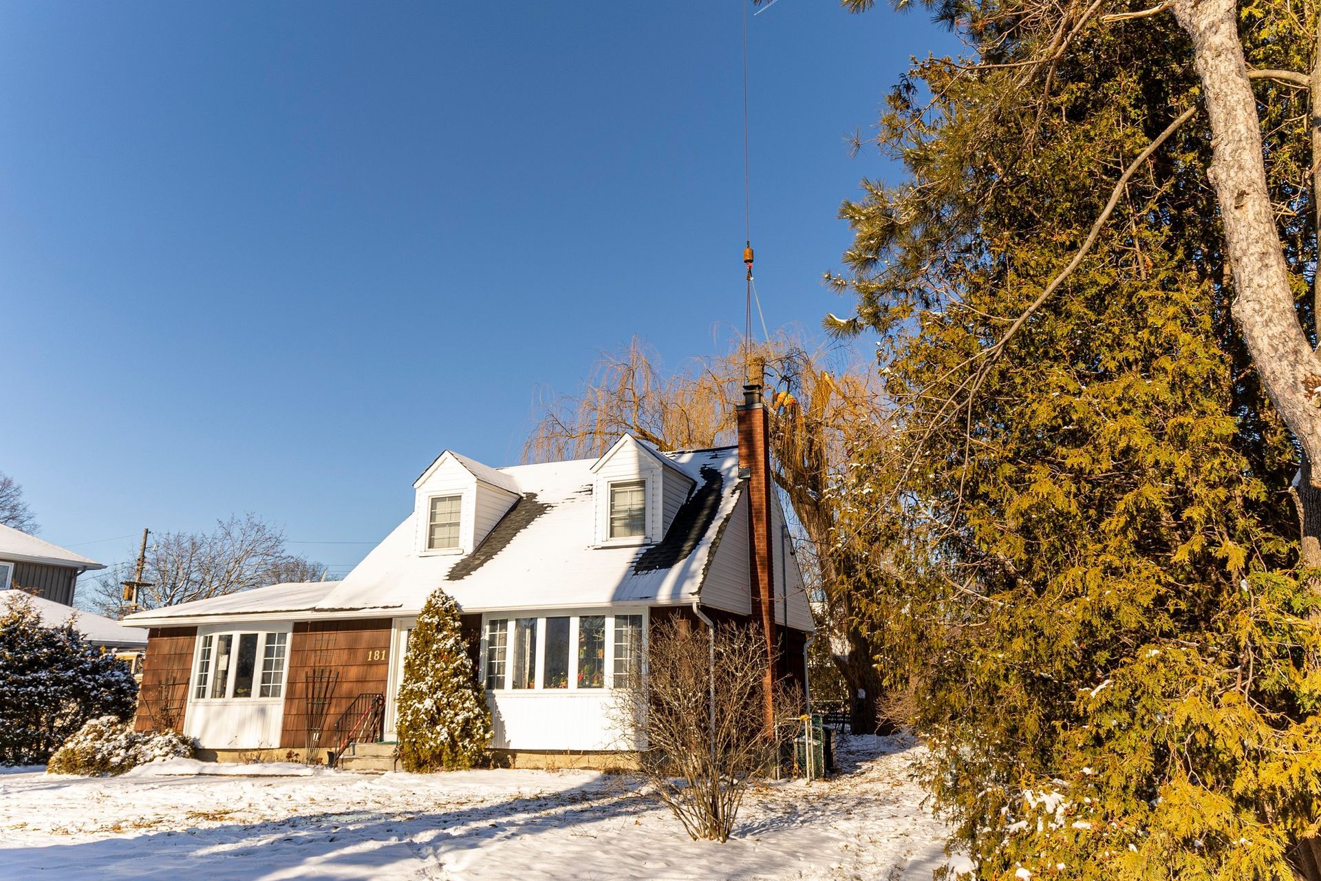 Snow-covered house with dormers on a sunny winter day. Tall trees to the right, clear blue sky.
