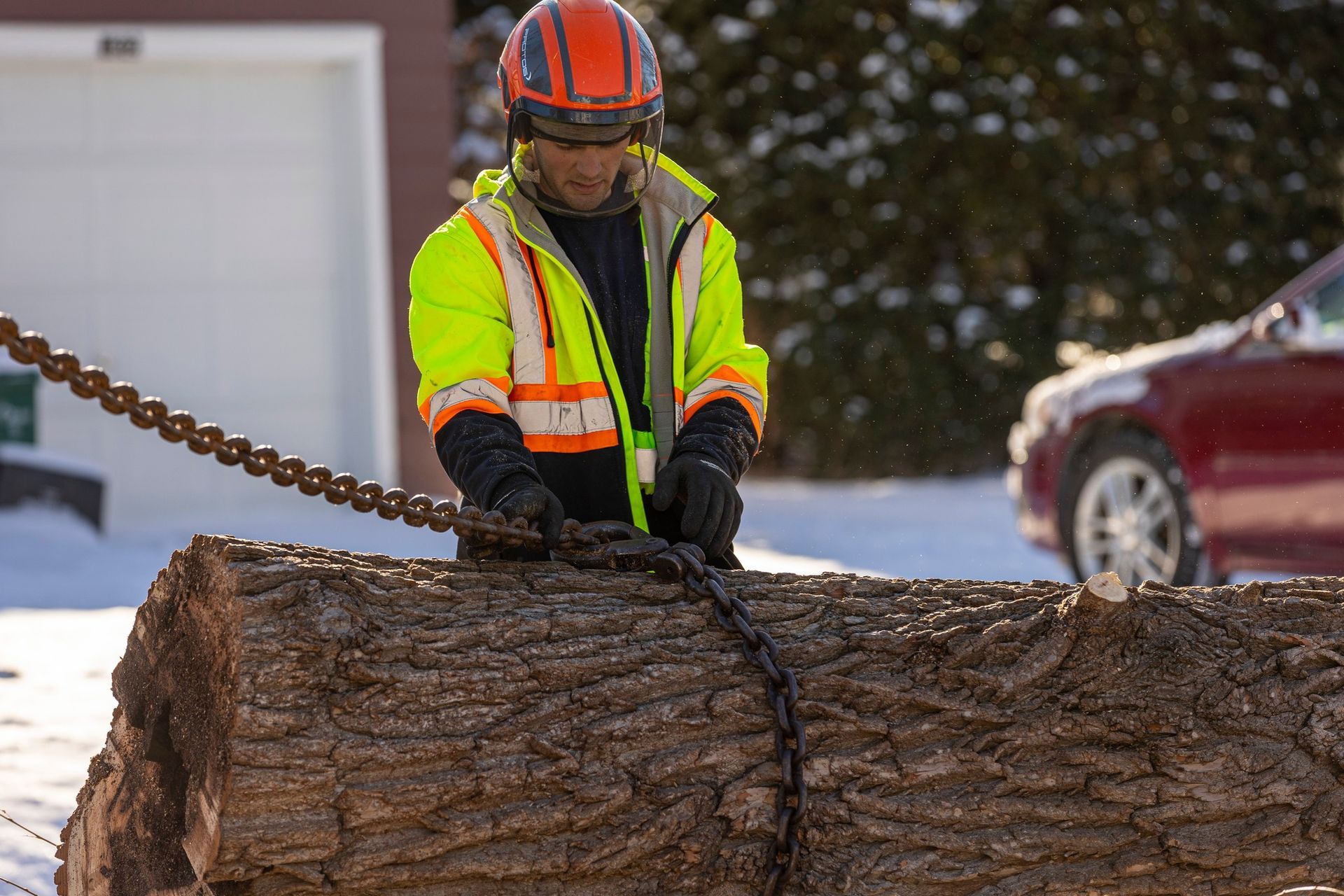 Lumberjack in high-vis vest secures a chain around a log in a snowy setting.