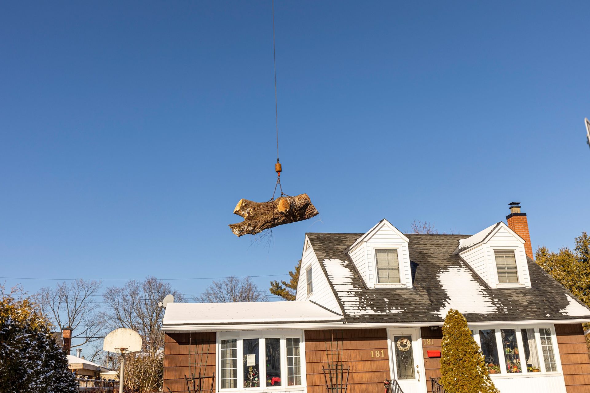 Crane lifting a large pile of tree branches in front of a house on a sunny day.