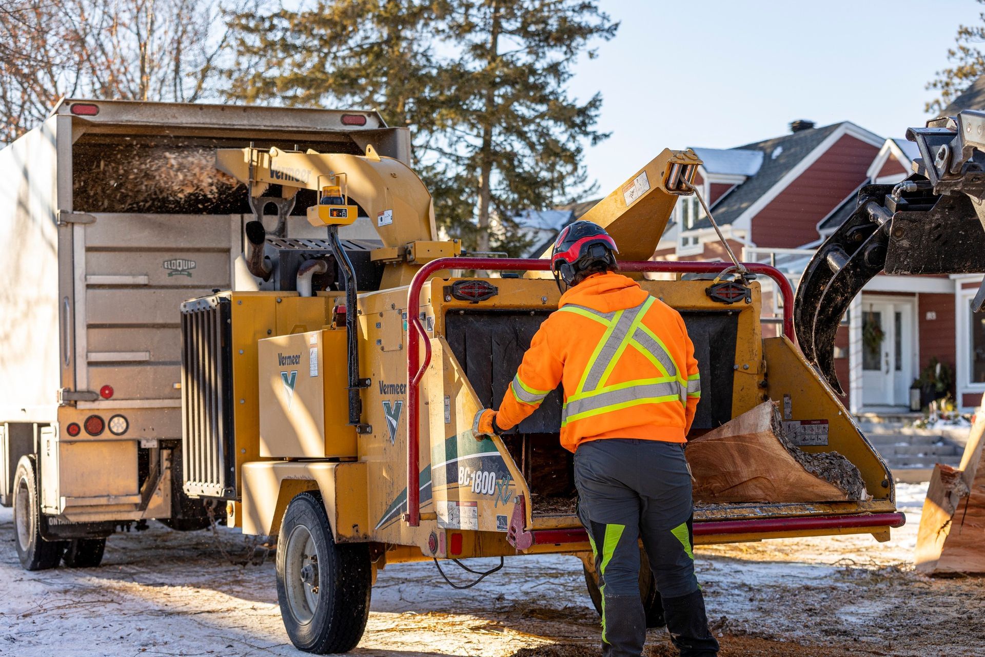 Man in orange vest feeds wood into a wood chipper next to a truck in a snowy residential area.