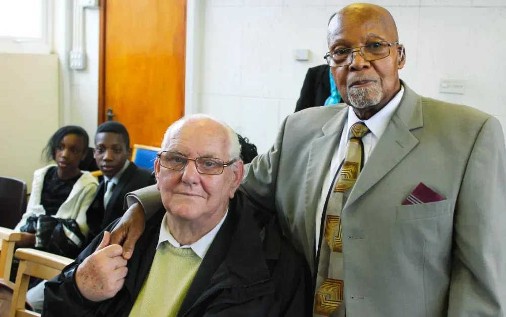 A man in a suit and tie is standing next to another man in a chair