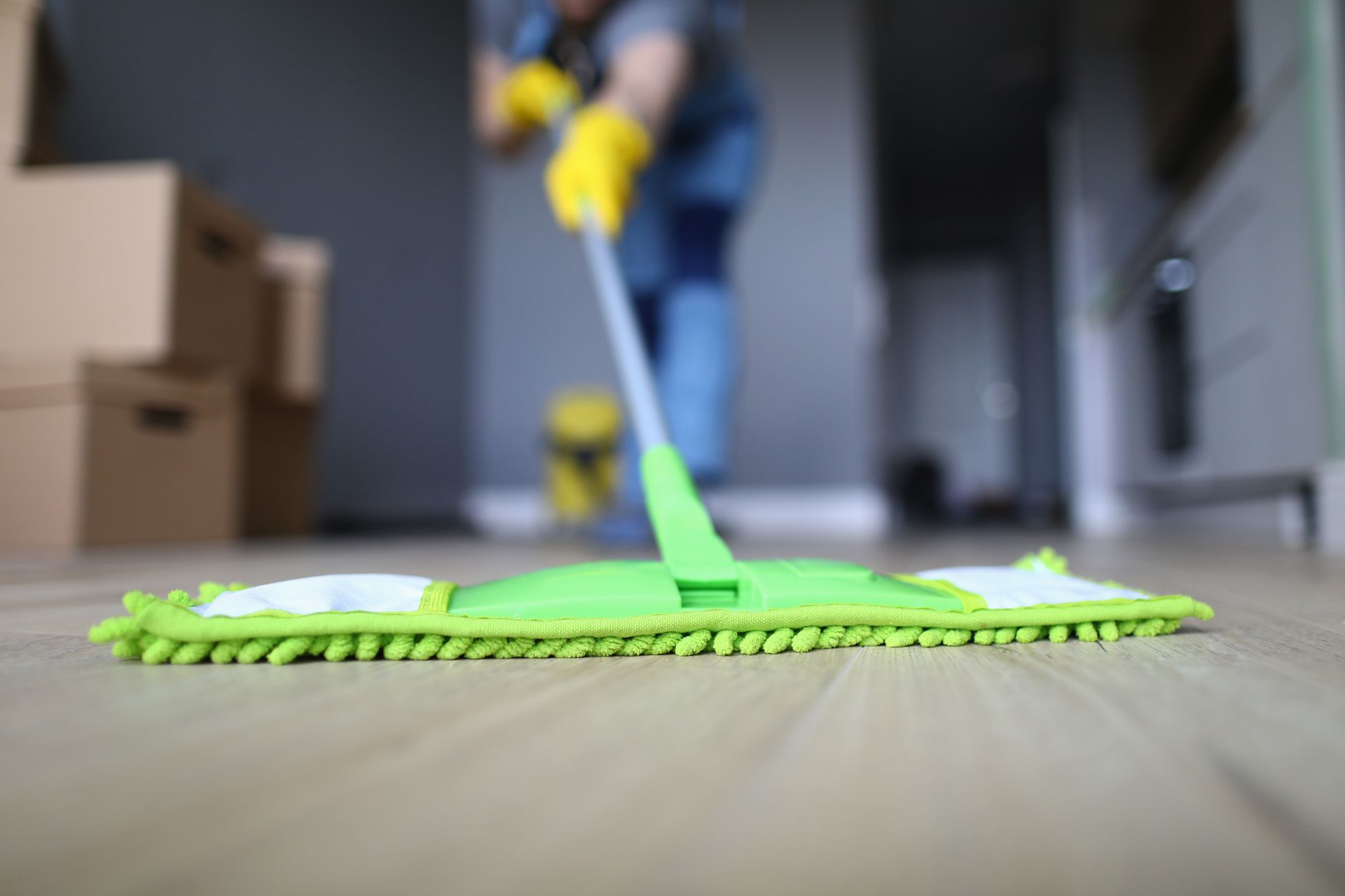 Person mopping a wooden floor with a green mop, wearing yellow gloves, inside a room with boxes.
