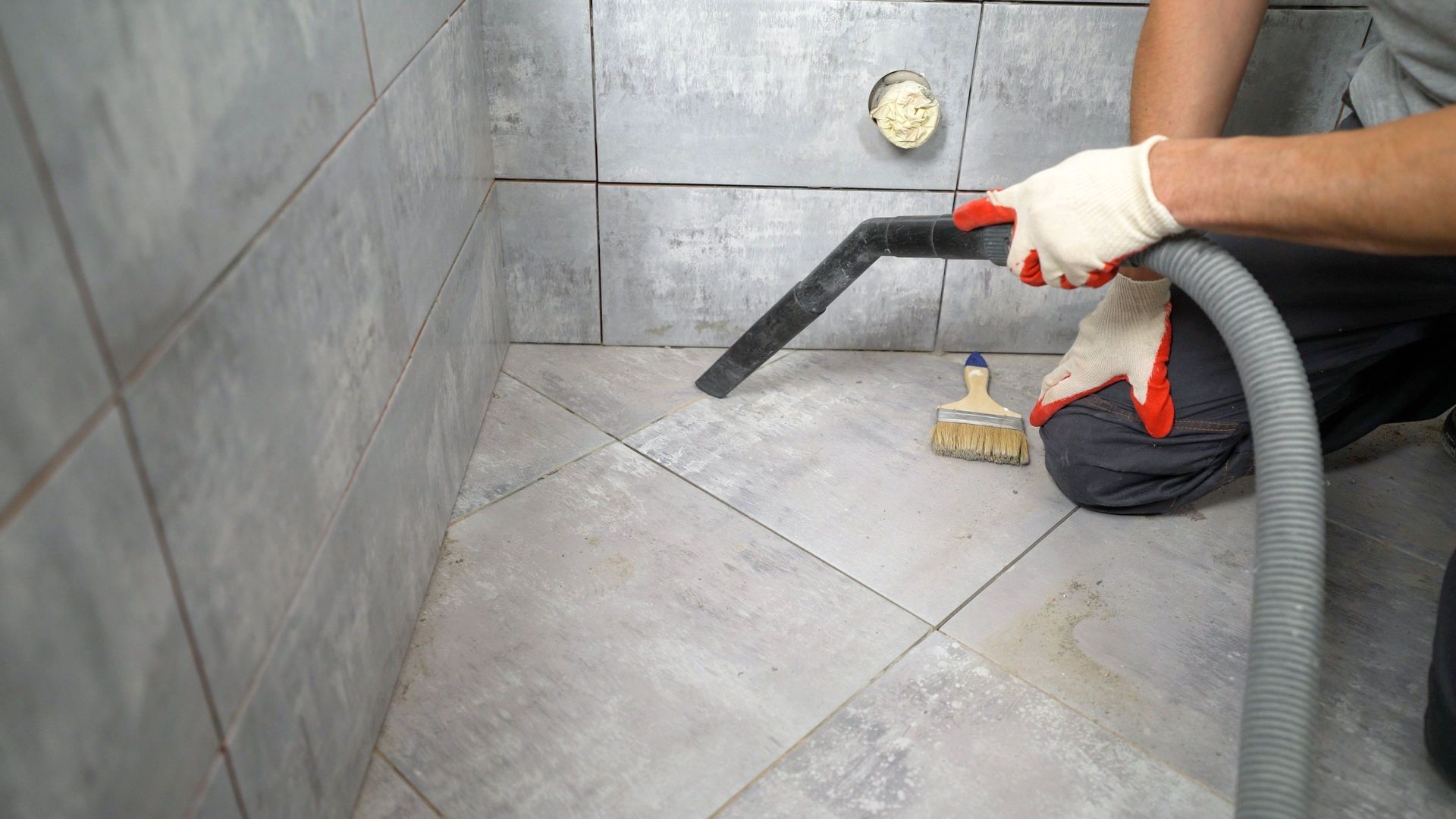 Person vacuuming grout from tiled shower floor with a brush nearby.