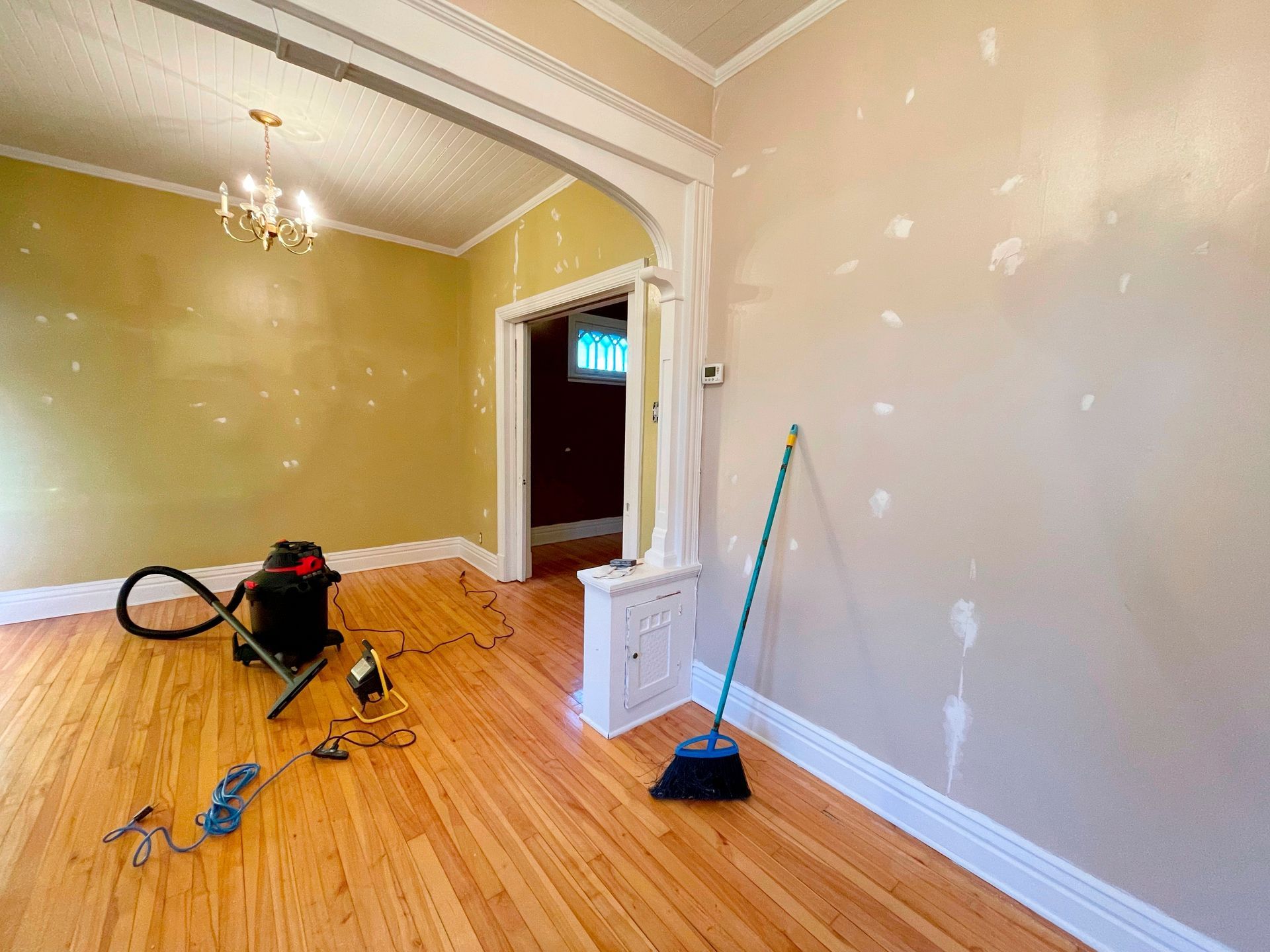 Interior room under renovation, hardwood floors, tan walls with patched spots, vacuum, tools, broom.