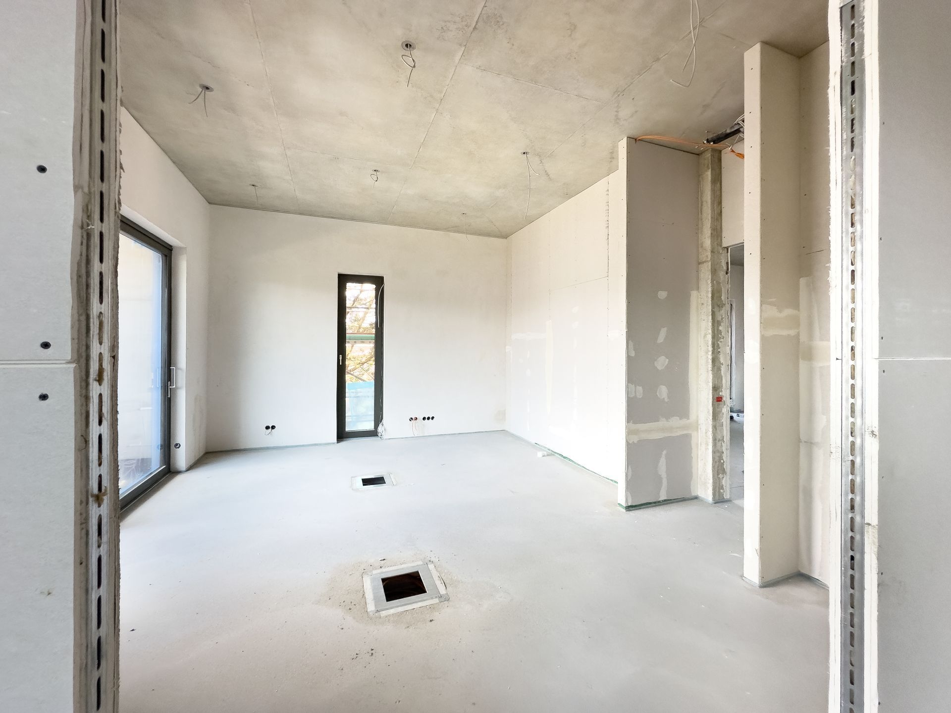 Interior of a room under construction, with drywall, window and a square hole in the floor.