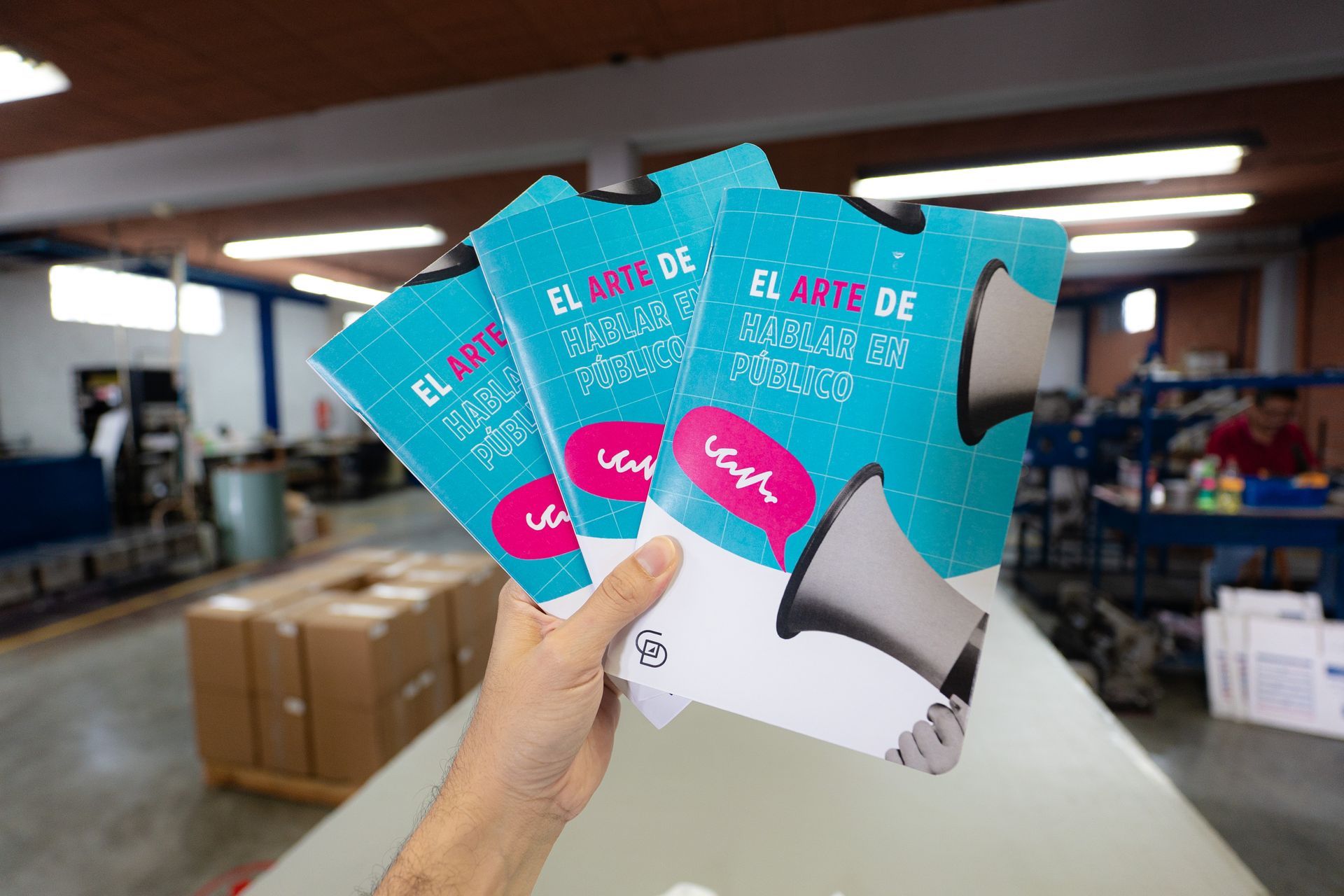 Hand holding three turquoise-and-white brochures in a print shop, with boxes and equipment in the background