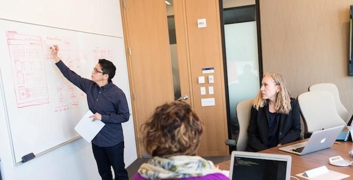 Tres personas colaboran en una reunión de oficina mientras una de ellas dibuja diagramas en una pizarra blanca.