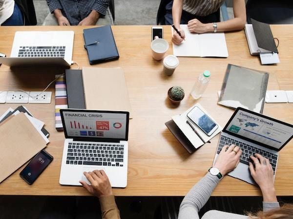 Personas trabajando alrededor de una mesa de madera con computadoras portátiles, cuadernos y café durante una reunión.