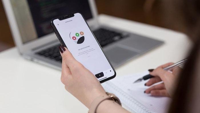 Primer plano de unas manos sosteniendo un teléfono inteligente con una aplicación abierta, con un ordenador portátil y un cuaderno al fondo.