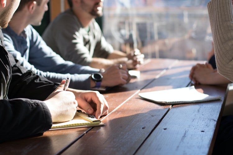 Un grupo de personas está sentado alrededor de una larga mesa de madera, escribiendo en libretas durante una reunión.