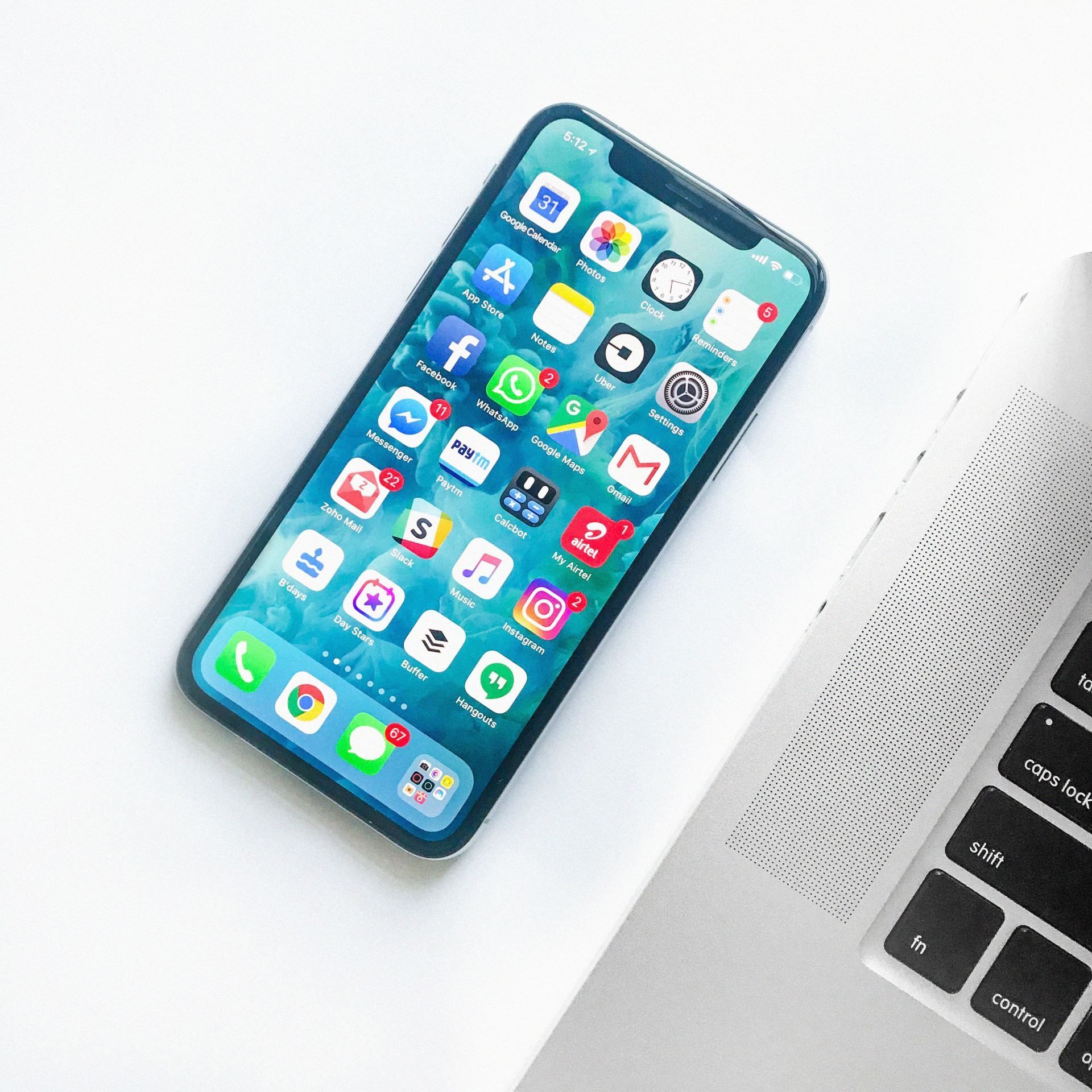 Un iPhone con varios iconos de aplicaciones en su pantalla, colocado junto a la esquina del teclado de un portátil sobre una superficie blanca.