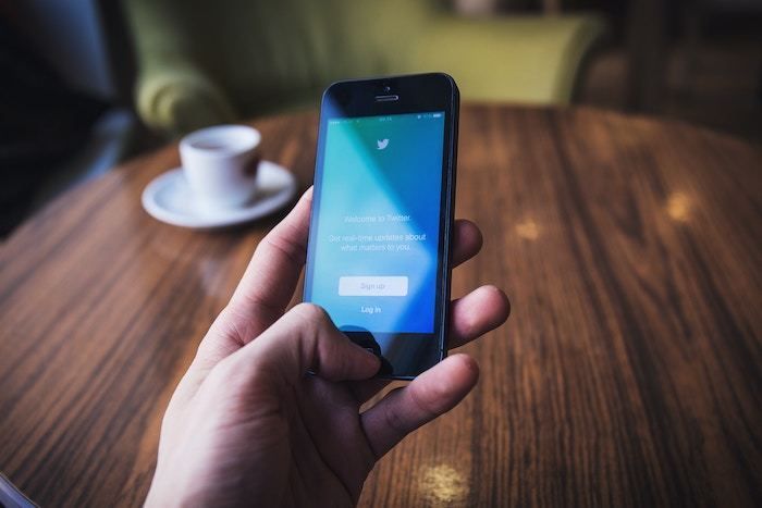 Primer plano de una mano que sostiene un teléfono inteligente con la pantalla de inicio de sesión de Twitter, sobre una mesa de madera con una taza de café.