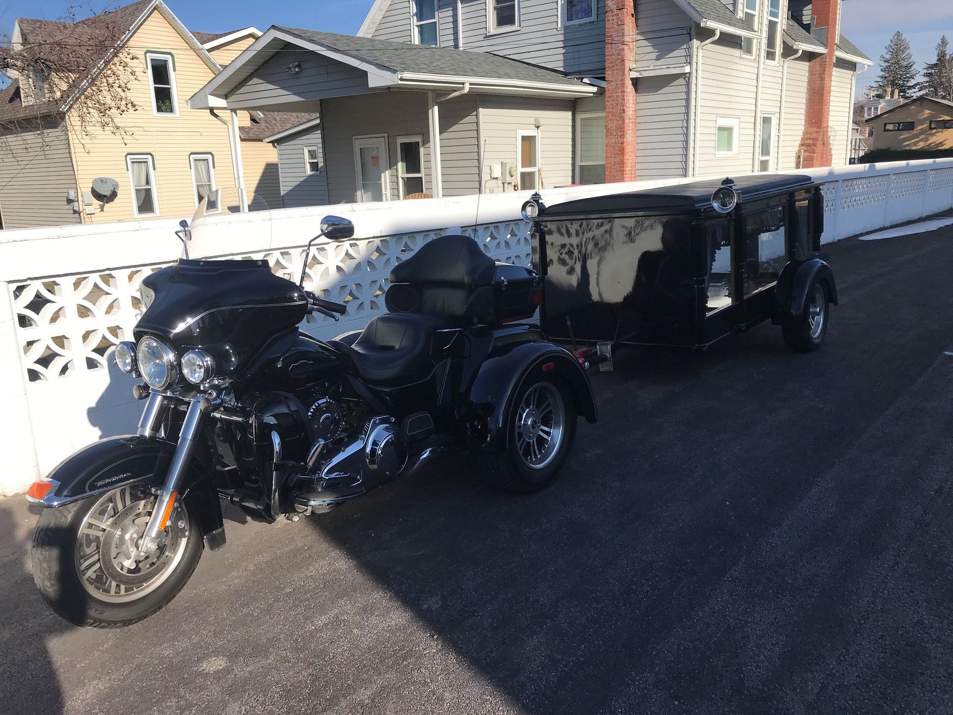 Black motorcycle trike pulling a hearse trailer in front of a house on a sunny day.