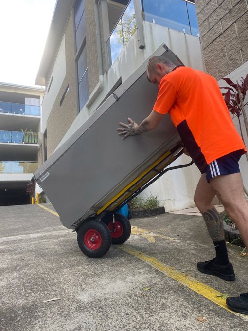 A Man In An Orange Shirt Is Pushing A Cart With A Large Box On It — Uplift Removals in Burdell, QLD