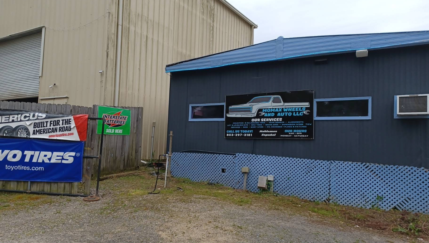 Small auto shop exterior with blue trim, tire sign, and parked building signs on a gravel lot. | Momax Wheels and Auto LLC