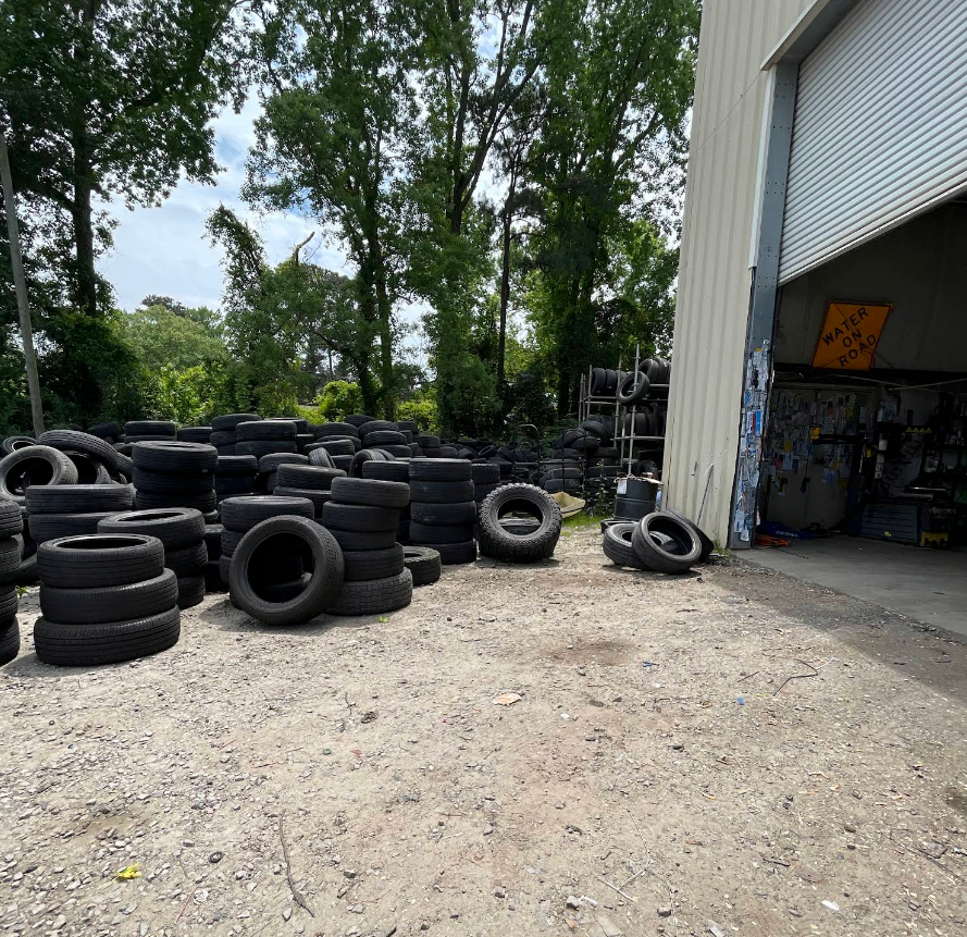 Stacks of used tires piled outside an open garage in a gravel lot, with trees in the background. | Momax Wheels and Auto LLC