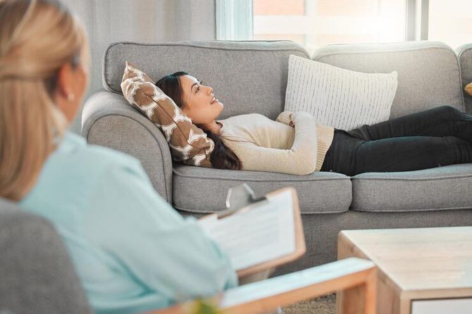 Person lying on a gray couch during a therapy session, speaking with a seated counselor nearby