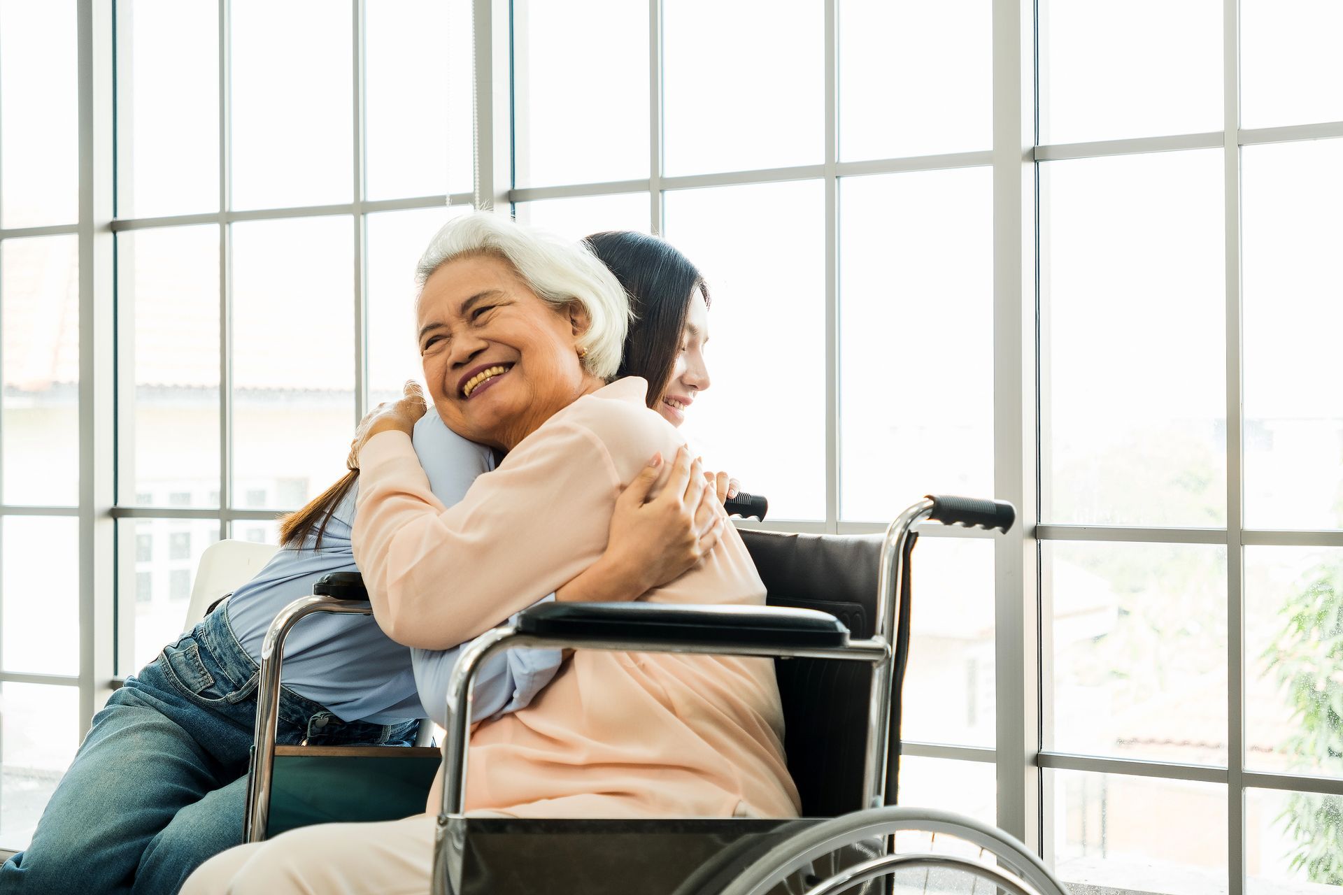 Woman in wheelchair embraced by another woman near a large window, smiling.