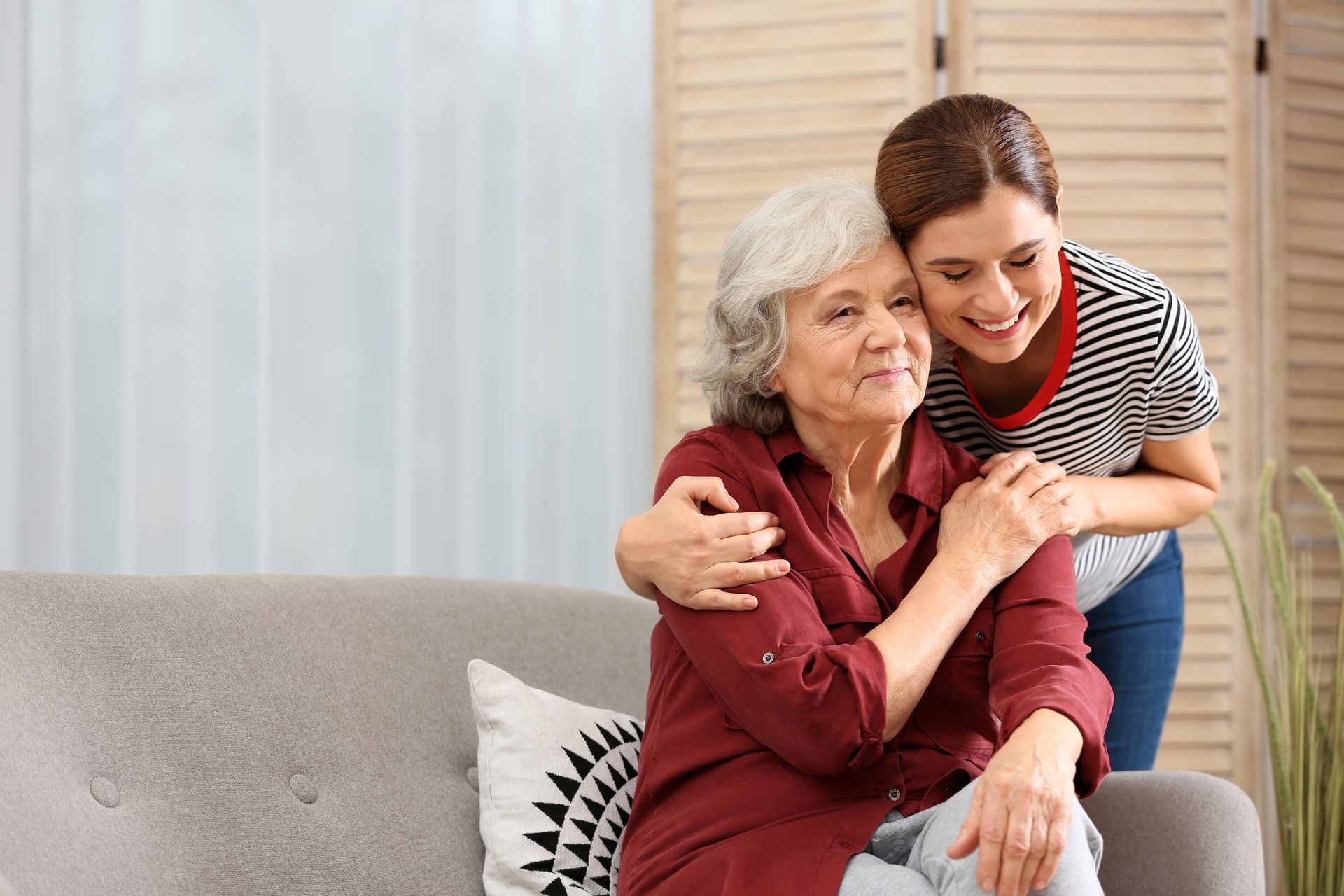Young woman hugs older woman seated on a sofa indoors. Both smile. Young woman hugs older woman seated on a sofa indoors. Both smile.