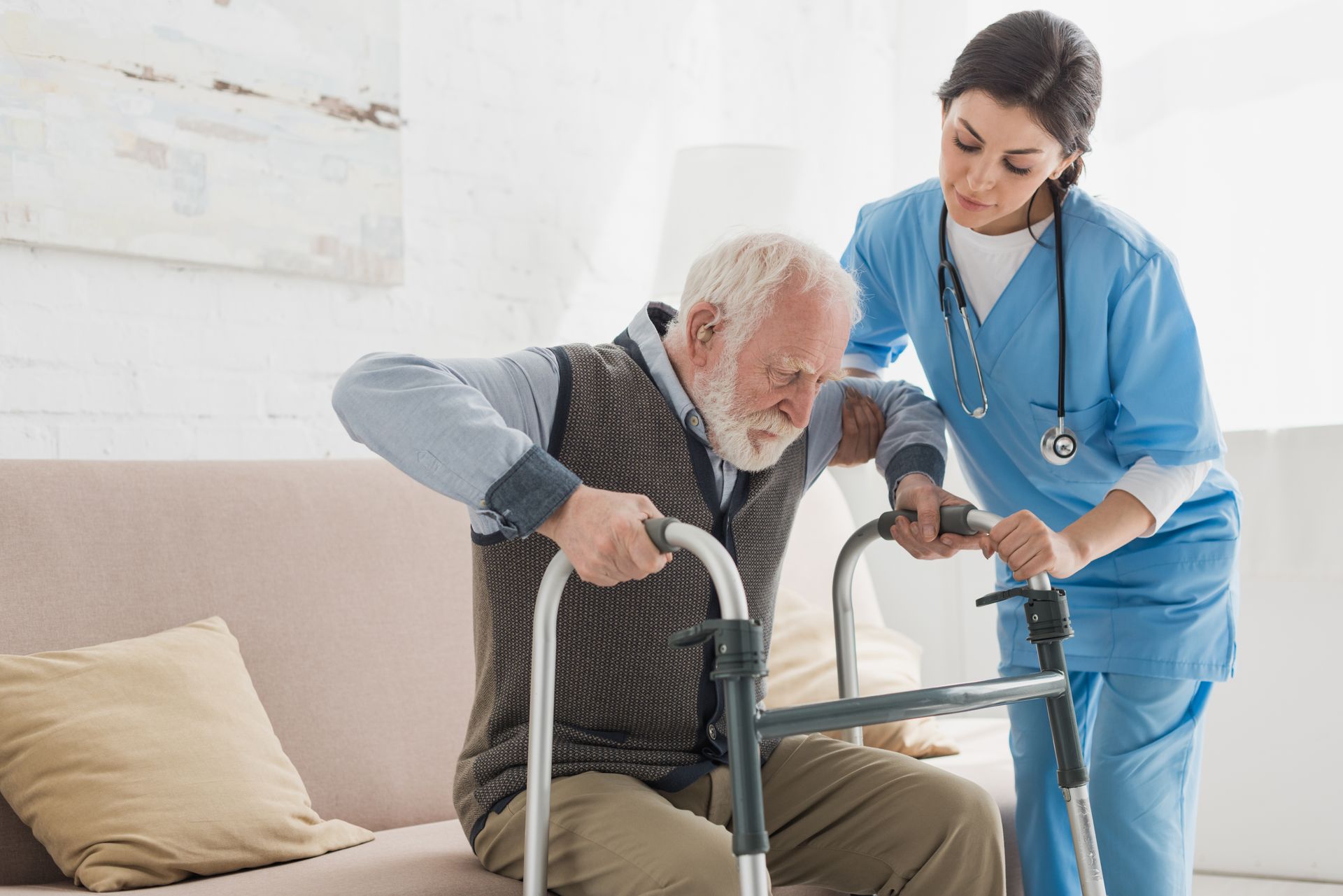 Nurse assists a man using a walker to stand from a couch.