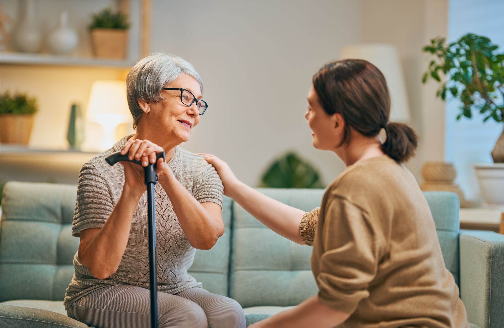 Woman with cane smiles, talking to another woman who has her hand on her shoulder; they sit on a sofa.