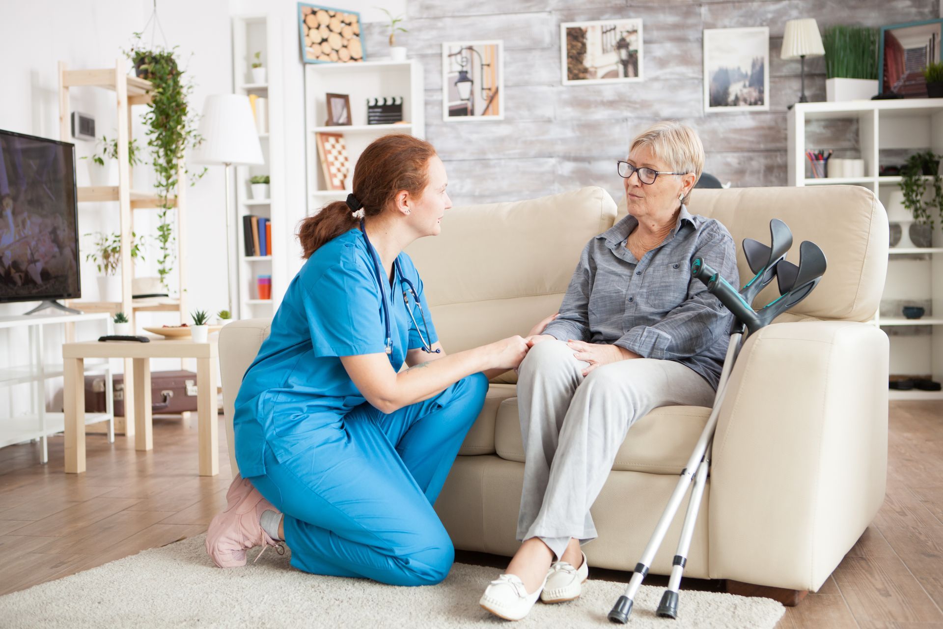 Woman in blue scrubs kneels, holding hand of older woman on sofa with crutches nearby.
