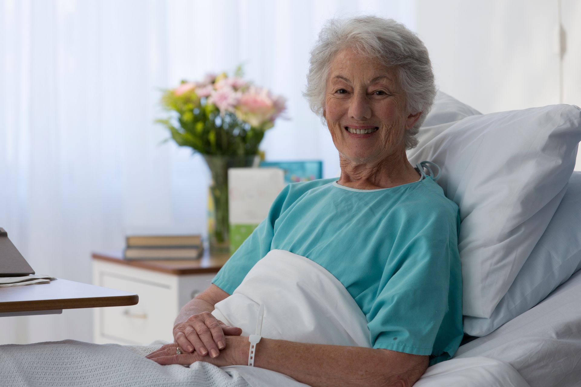 Smiling person in hospital bed, wearing teal gown. Flowers and books on bedside table.