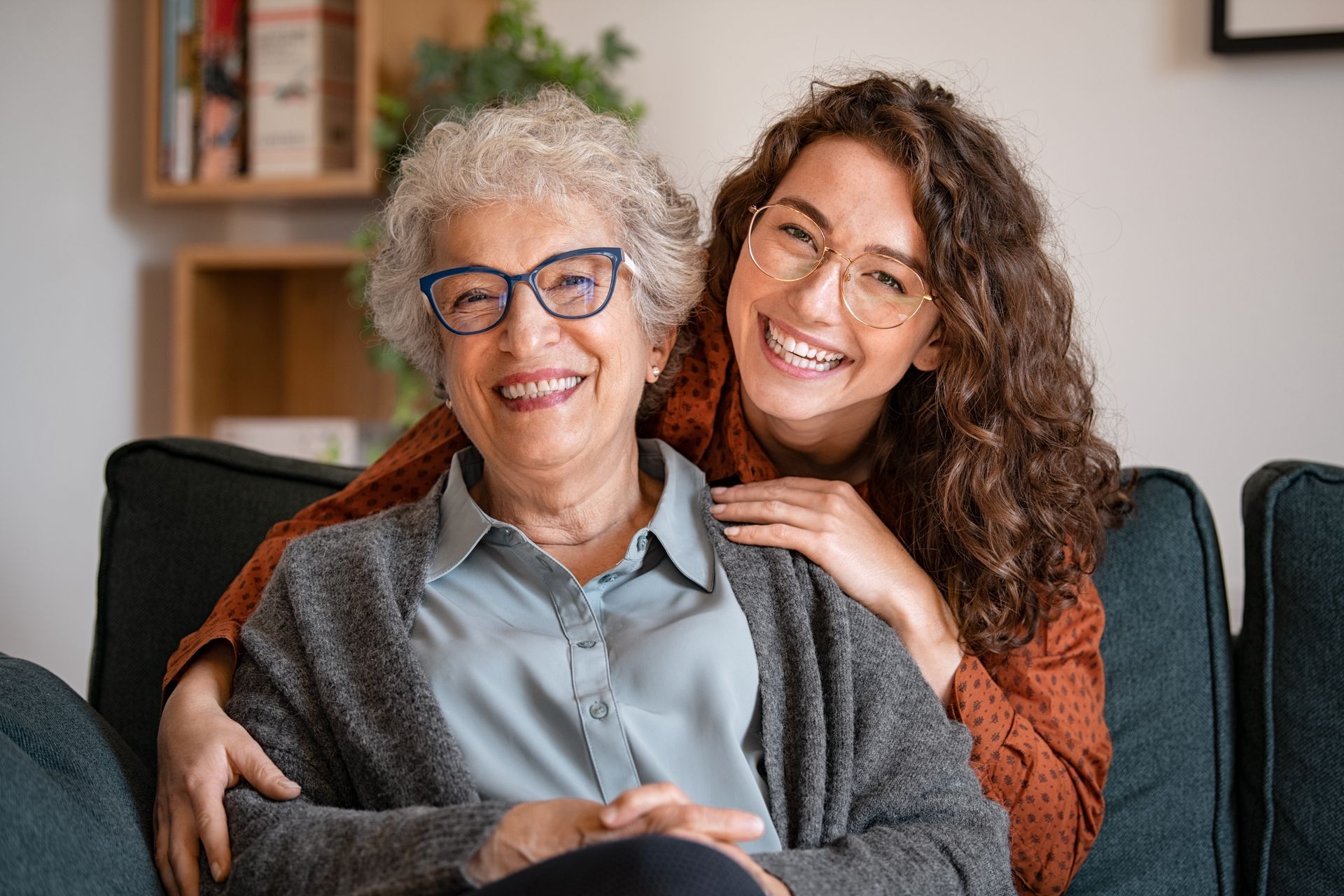 Woman with curly hair embraces elderly woman with glasses, both smiling on a sofa.