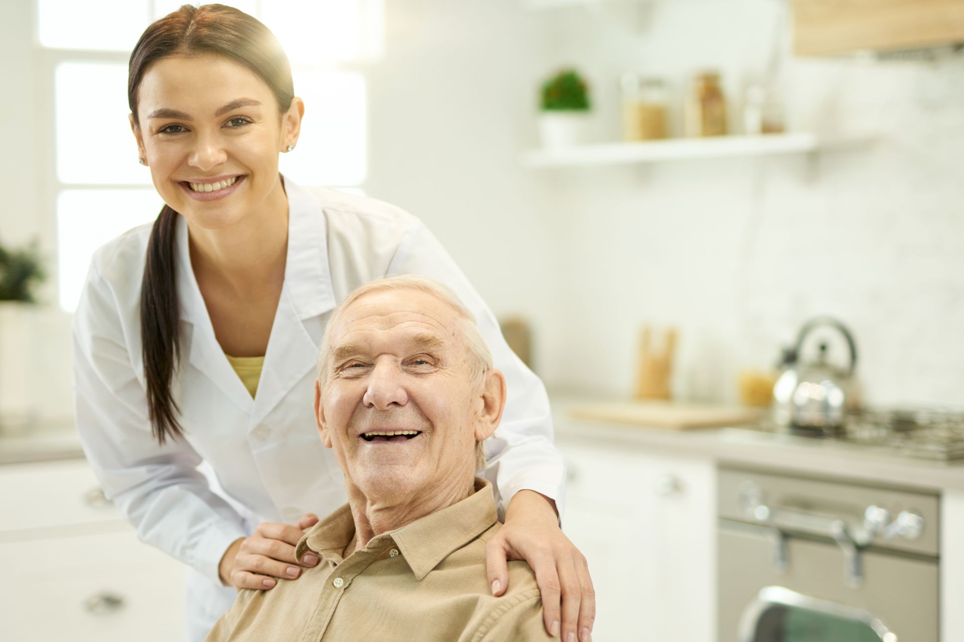 Woman in white coat smiles with arm on an older man's shoulder in a kitchen.
