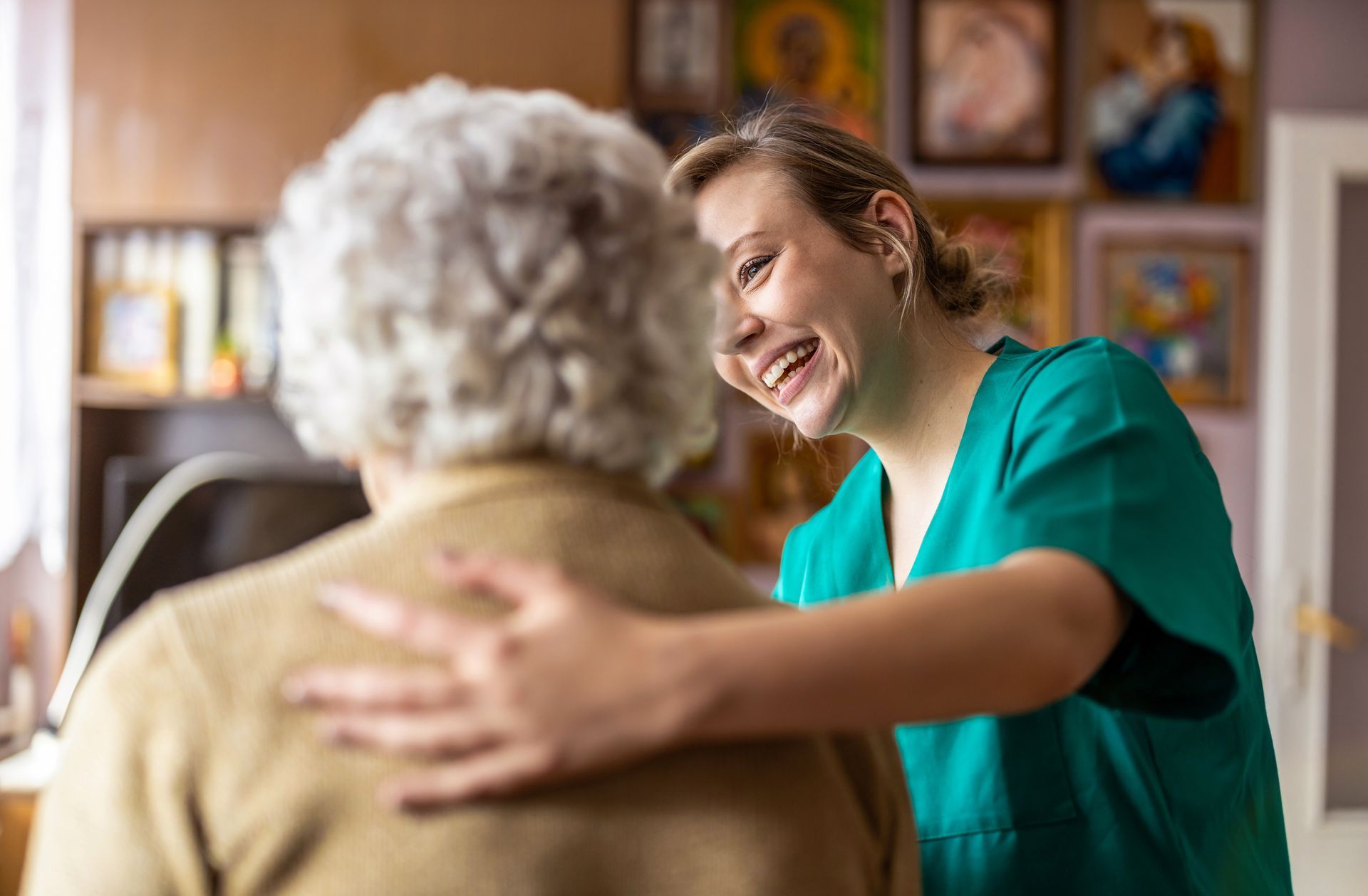 A smiling person in teal scrubs comforts an older person, indoors with paintings.