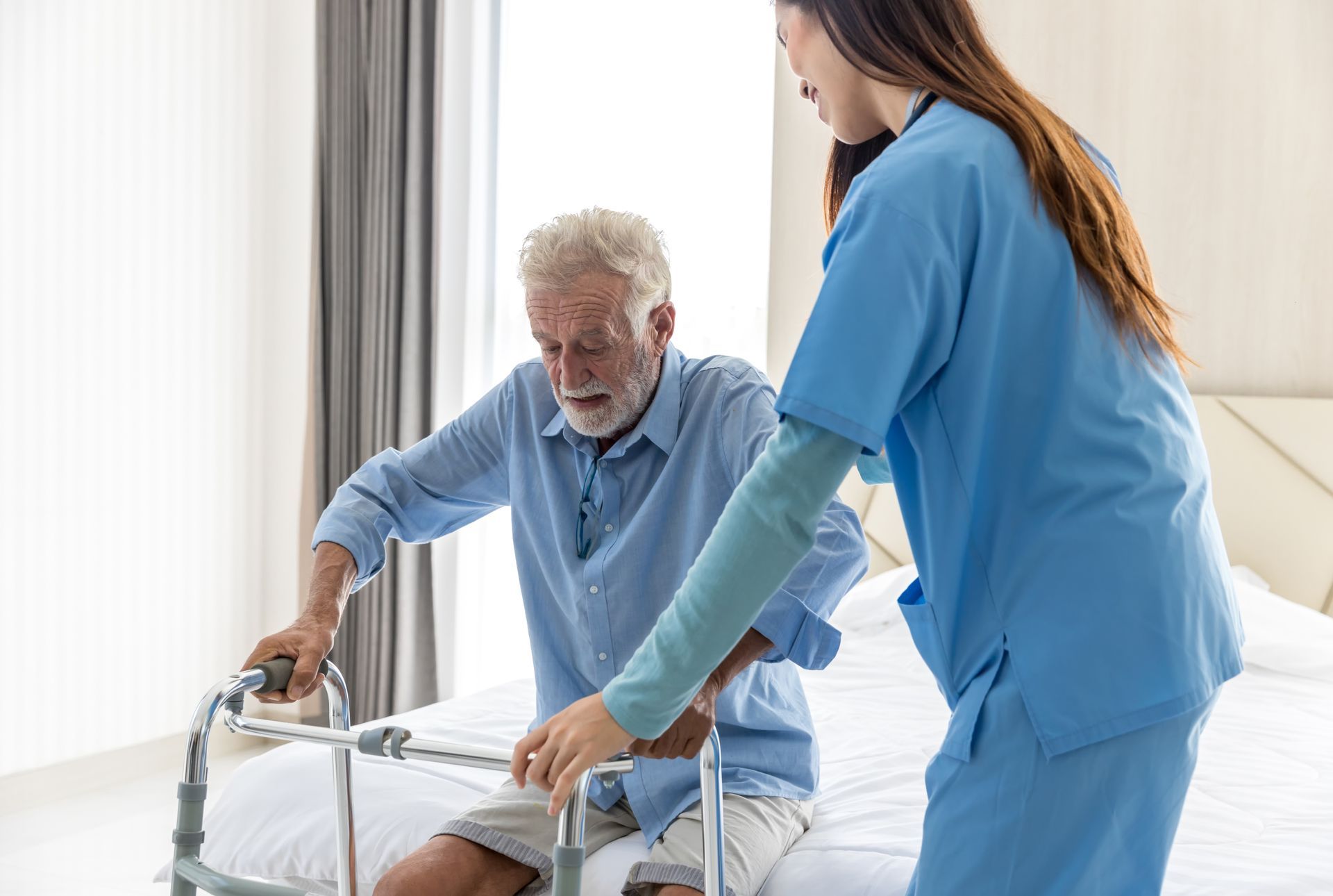 Nurse assisting a person using a walker to stand up from a bed in a room.