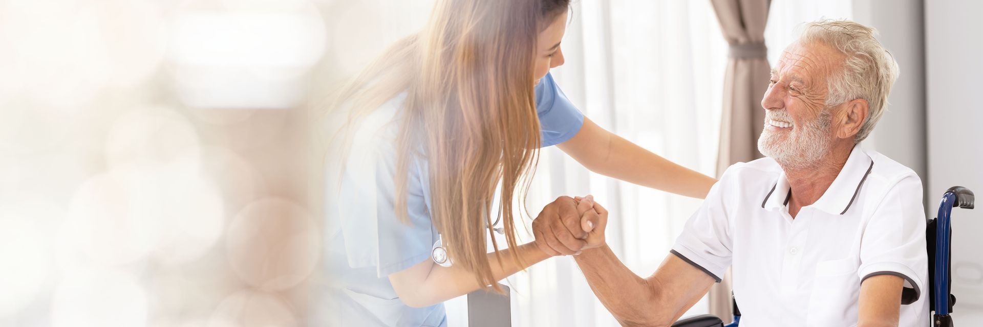 Caregiver assisting a person in a wheelchair. They hold hands and smile indoors near a window.