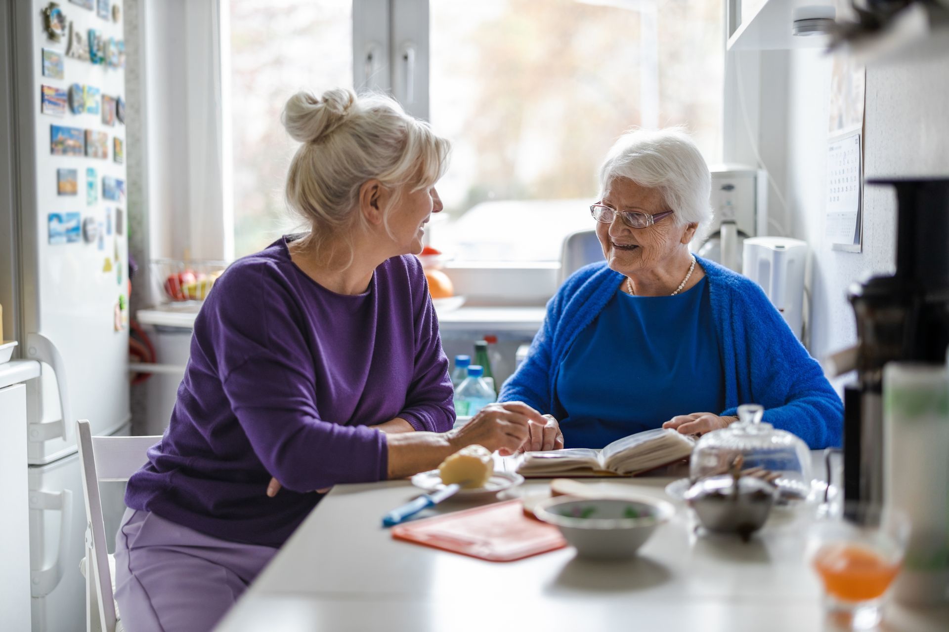 Two women in a kitchen, one wearing purple, one in blue, looking at a book together at a table. Two women in a kitchen, one wearing purple, one in blue, looking at a book together at a table.