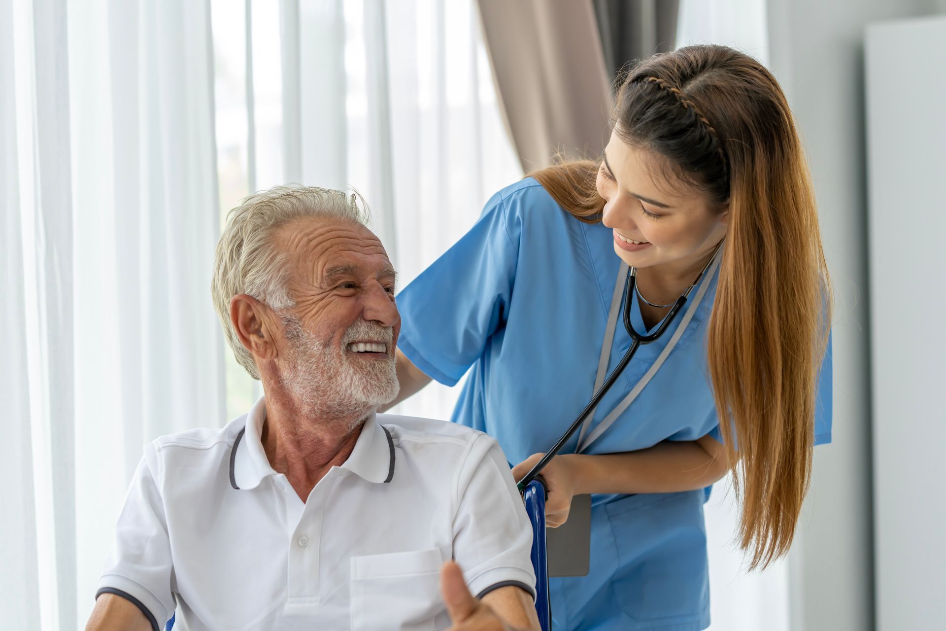 Nurse assisting an elderly person seated in a wheelchair, both smiling, inside a home.