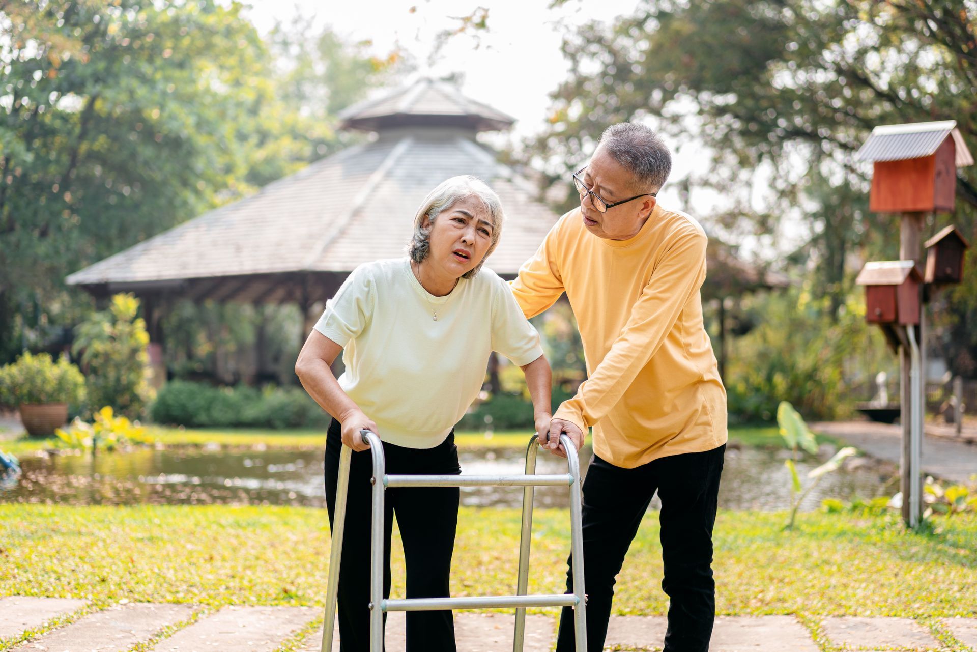Elderly person using walker, assisted by another, in a park setting.